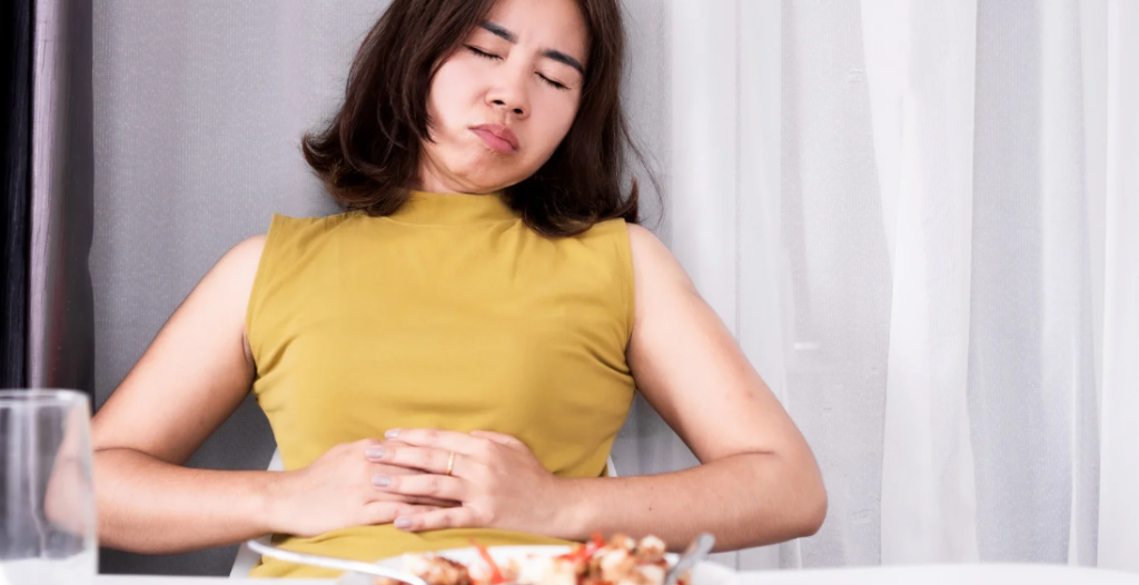 Mujer sentada con las manos en su abdomen y con los ojos cerrados frente a un plato de comida porque tiene inflamación abdominal